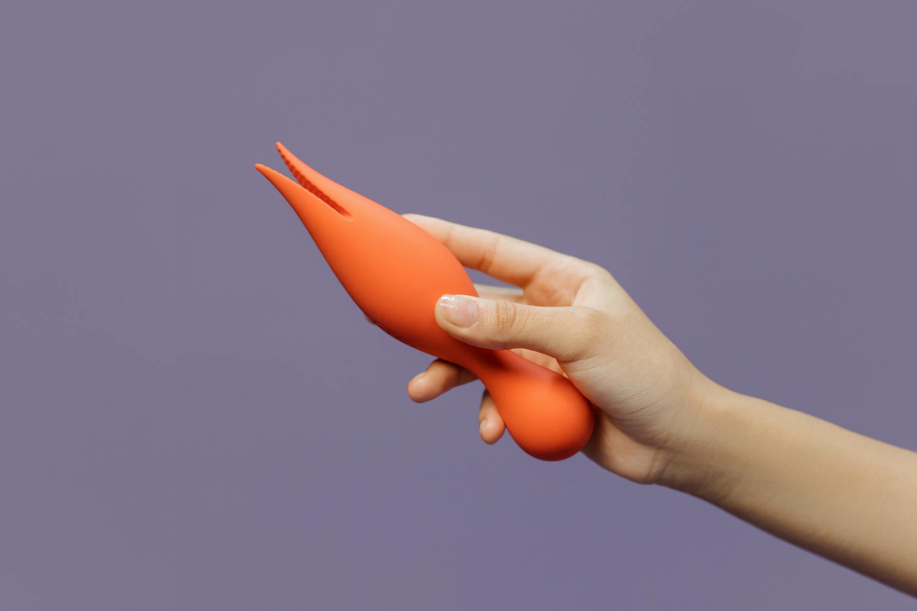 A hand holding an orange vibrator against a minimalistic purple backdrop, showcasing modern sensuality and intimate exploration.